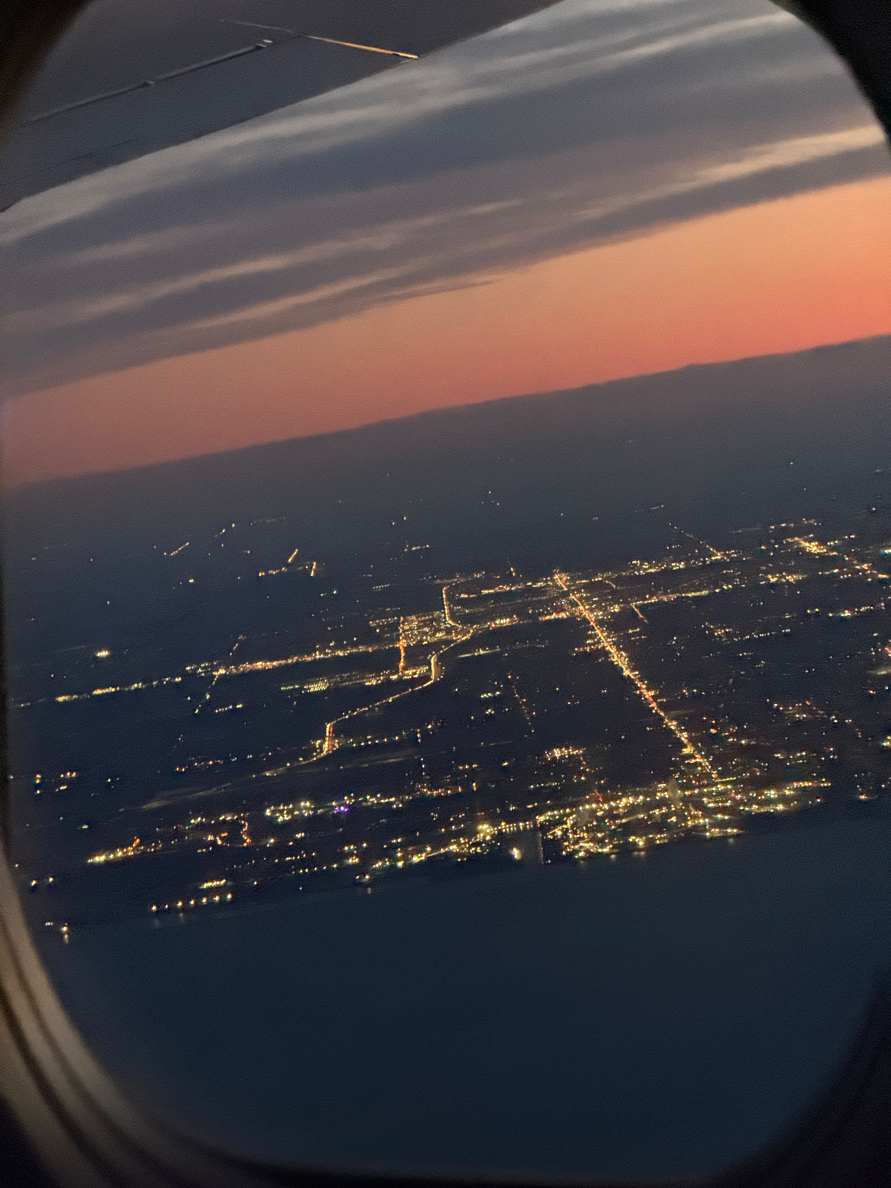 An aerial view from an airplane window shows a city illuminated with lights beneath a darkening sky at sunset.